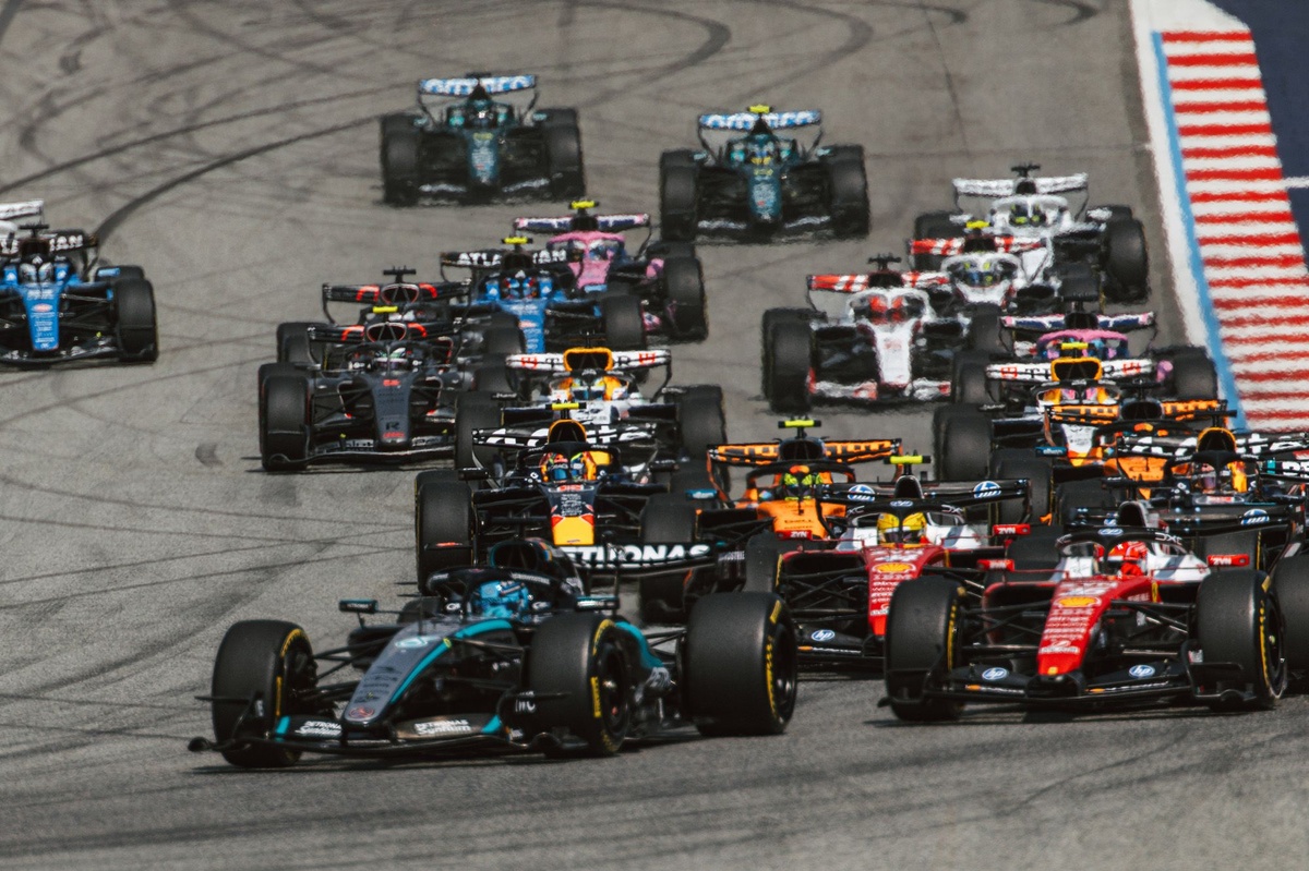 A dramatic wide shot of a Formula 1 car speeding down a track under intense stadium lights, with a blurred Brazilian flag subtly visible in the background.